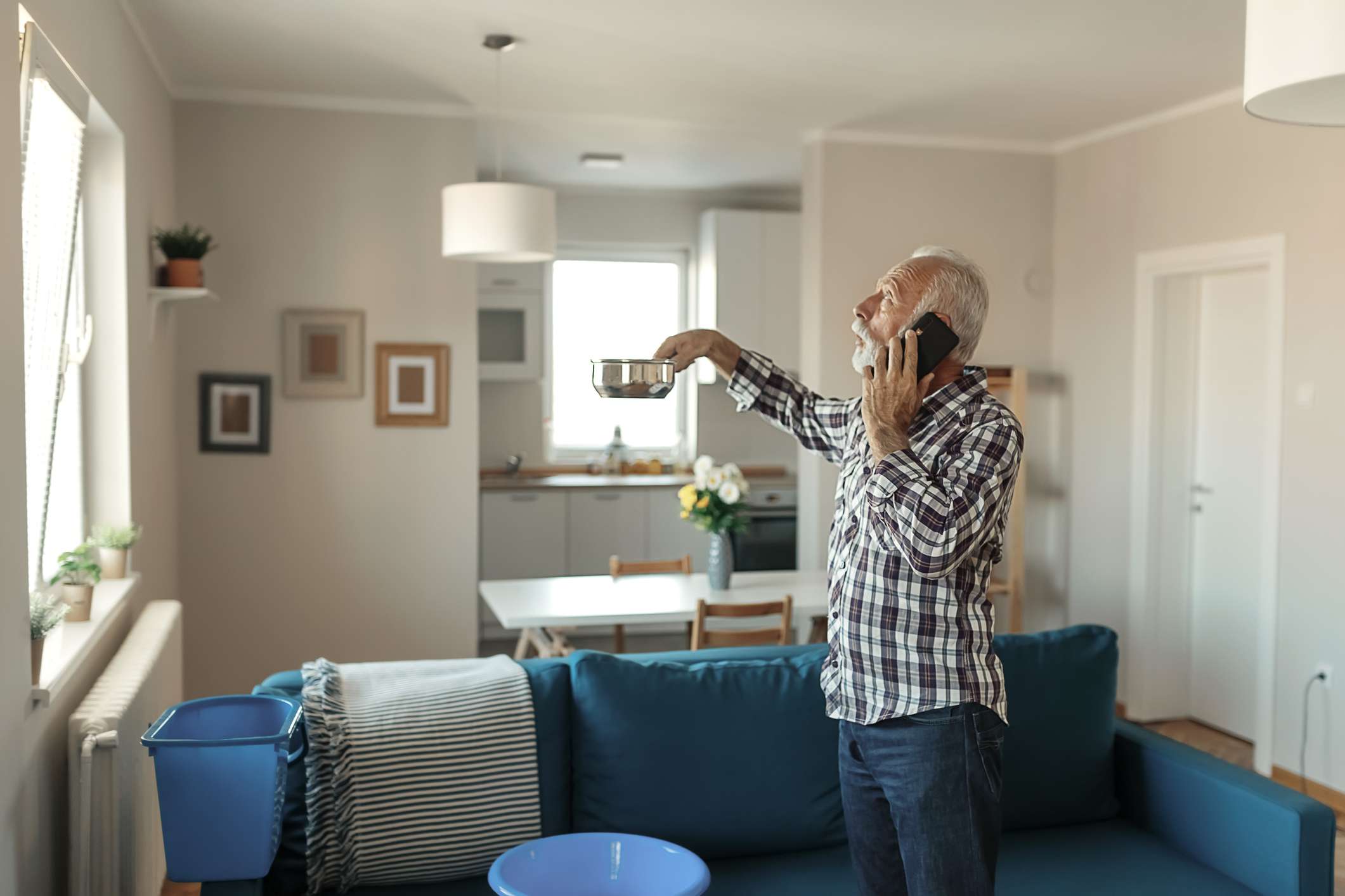 Skäggig äldre man som talar på smartphonen nära plast- tvättbunke hemma i vardagsrummet på grund av läckande av tak